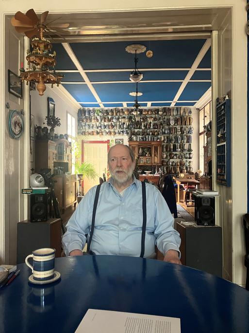 Ein Mann sitzt an einem Tisch, hinter ihm an der Wand ist eine Keramiksammlung sichtbar