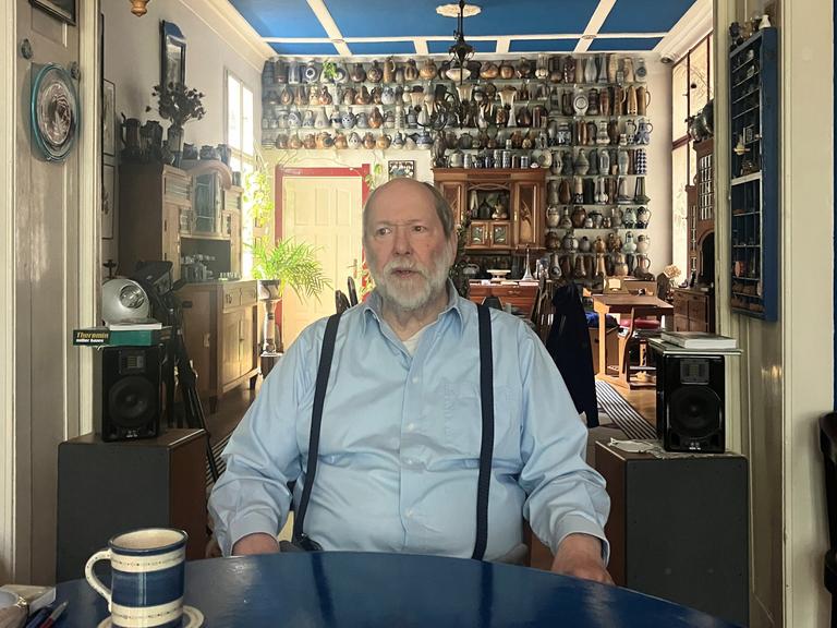 Ein Mann sitzt an einem Tisch, hinter ihm an der Wand ist eine Keramiksammlung sichtbar