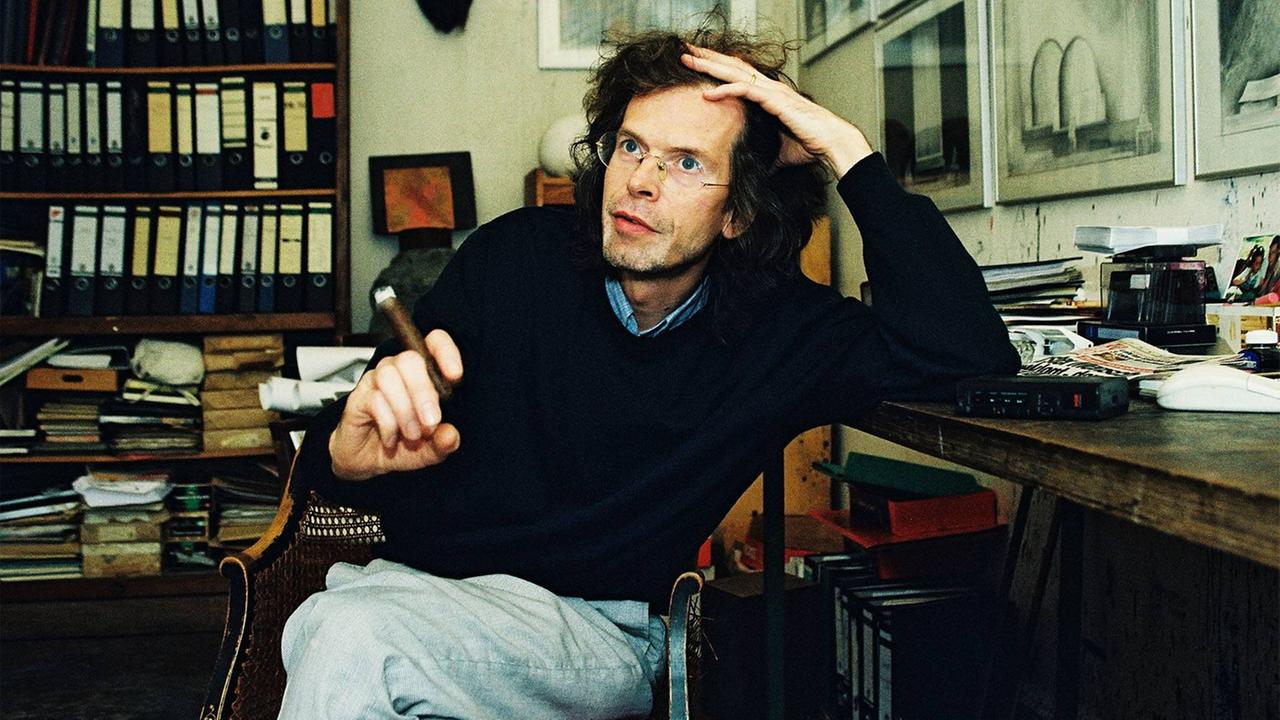 Florian Havemann sitzt mit einer Zigarre an einem Schreibtisch in einer Wohnung voller Bücherregale