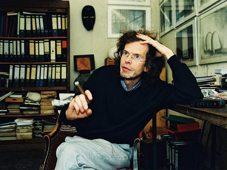Florian Havemann sitzt mit einer Zigarre an einem Schreibtisch in einer Wohnung voller Bücherregale Florian Havemann sitzt mit einer Zigarre an einem Schreibtisch in einer Wohnung voller Bücherregale