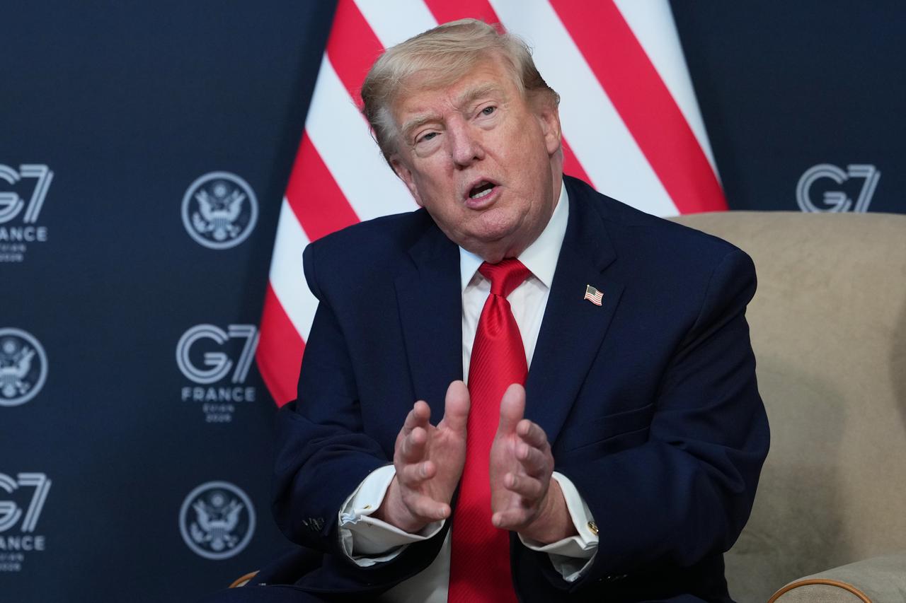 Donald Trump sitzt auf einem Sessel und gestikuliert mit den Händen. Im Hintergrund eine Flagge der Vereinigten Staaten.