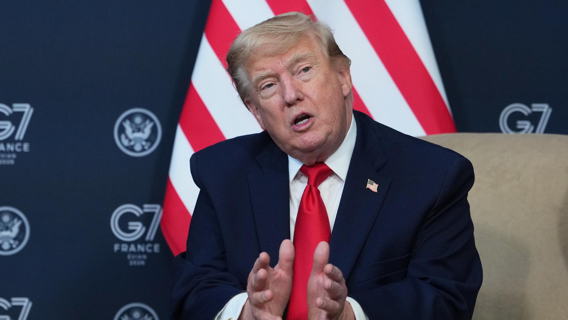Donald Trump sitzt auf einem Sessel und gestikuliert mit den Händen. Im Hintergrund eine Flagge der Vereinigten Staaten.