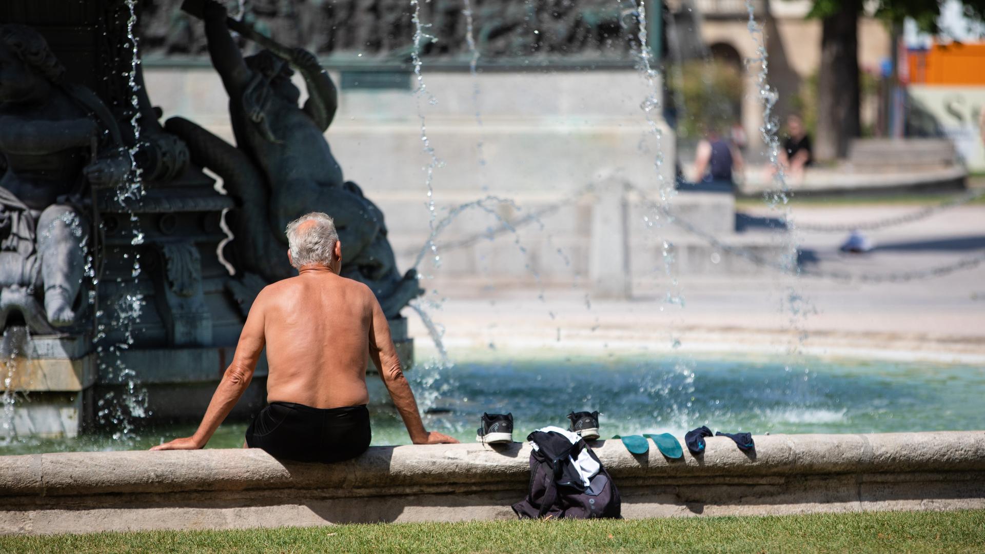 Ein älterer Mann sitzt bei heißen Temperaturen oben ohne auf dem Rand eines Brunnens, seine Kleidung liegt neben ihm.