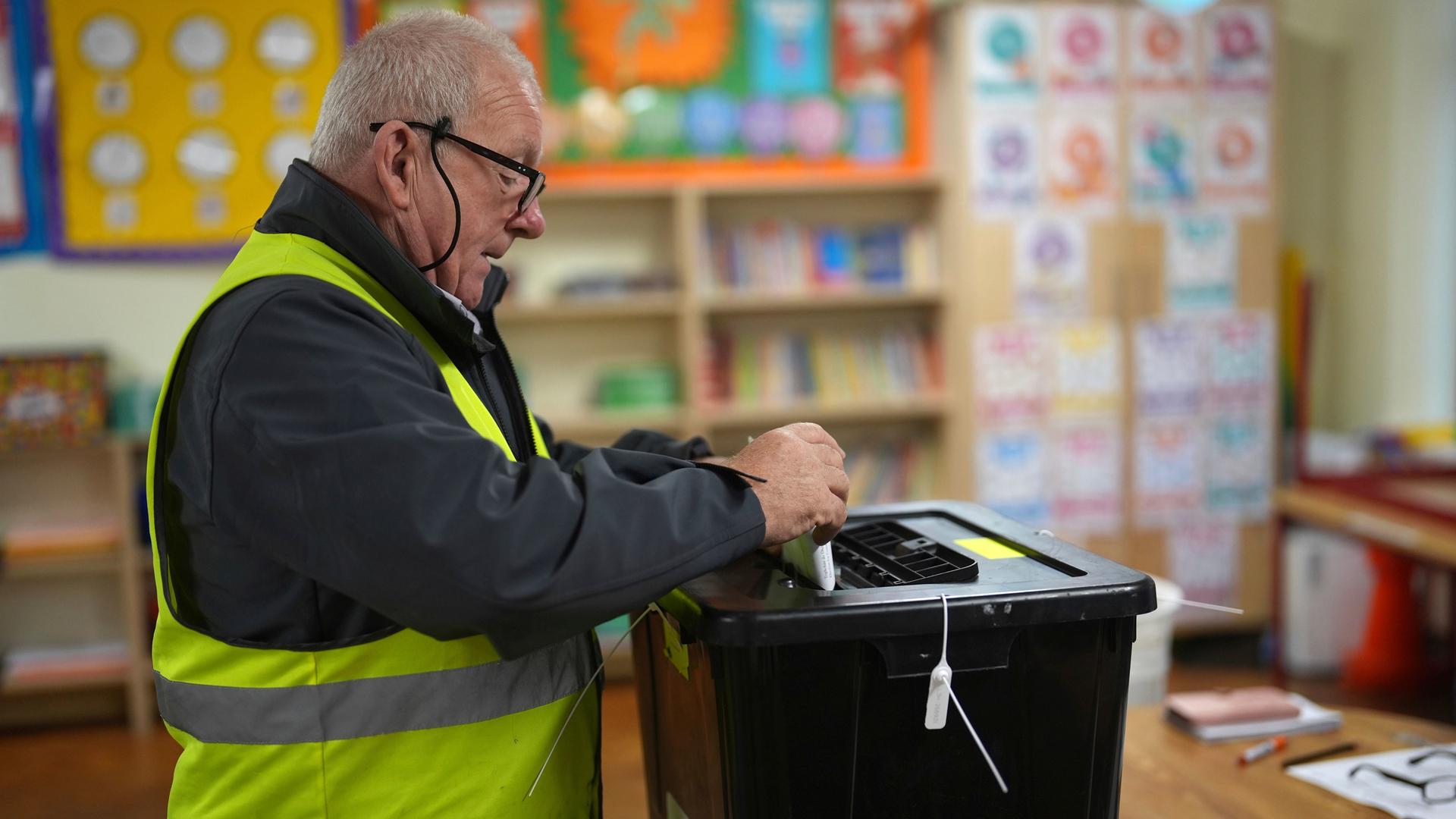 Der ältere Mann trägt eine gelbe Warnweste und eine Brille. Man sieht ihn von der Seite. Er steckt den Stimmzettel gerade in eine schwarze Wahlurne. Im Hintergrund unscharf gemalte Kinderbilder.