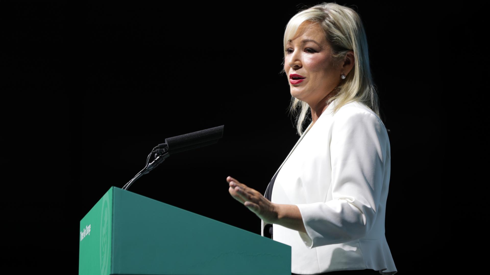 Man sieht sie von der Seite an einem Rednerpult sprechend. Sie trägt ein weißes Jackett und steht vor einem schwarzen Hintergrund. Ihre Haare sind blond und der Mund ist rot geschminkt.