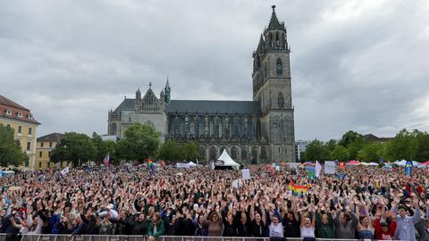 Tausende Menschen stehen auf dem Platz vor dem Magdeburger Dom und recken die Hände in die Höhe. Manche halten Plakate und Regenbogenfahnen in die Höhe.