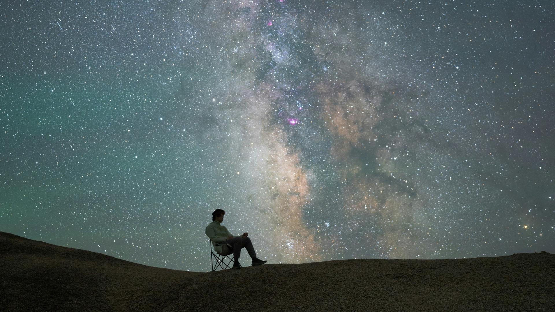 Ein Mann sitzt auf einem Klappstuhl auf einer Anhöhe und blickt in den wild funkelnden Sternenhimmel. Ein Mann sitzt auf einem Klappstuhl auf einer Anhöhe und blickt in den wild funkelnden Sternenhimmel.