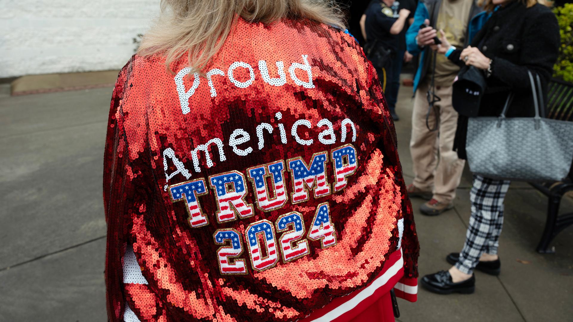 Eine blonde Donald-Trump-Wählerin trägt eine Glitzerjacke, auf der Rückseite steht: "PROUD AMERICAN - TRUMP 2024".