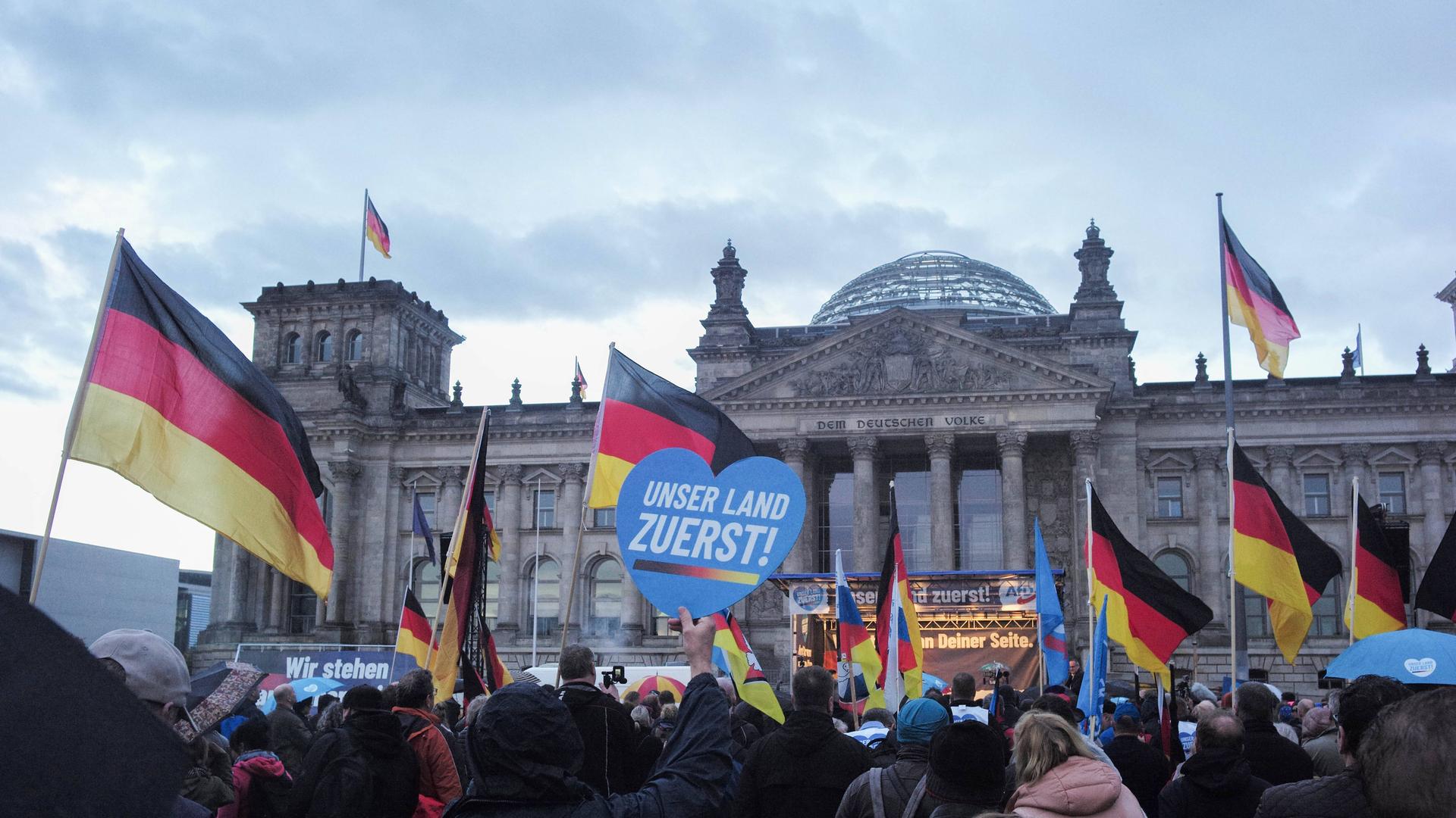 AfD-Anhänger und weitere Demonstrantinnen und Demonstranten vor dem Reichstagsgebäude in Berlin schwenken Deutschlandfahnen und ein blaues Herz mit der Aufschrift "Unser Land zuerst!"