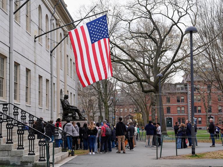 Besucher stehen unter der US-Flagge an der Statue von John Harvard an der Harvard Universität in Cambridge in den USA. 