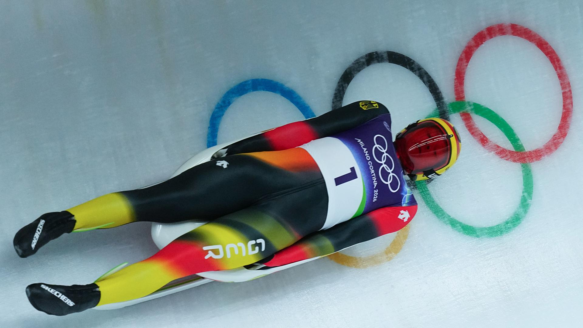 Der Rodler Max Langenhan liegt auf dem Rodelschlitten. Er trägt einen Anzug mit den deutschen Nationalfarben. Auf der Eisbahn sind die olympischen Ringe zu sehen.
