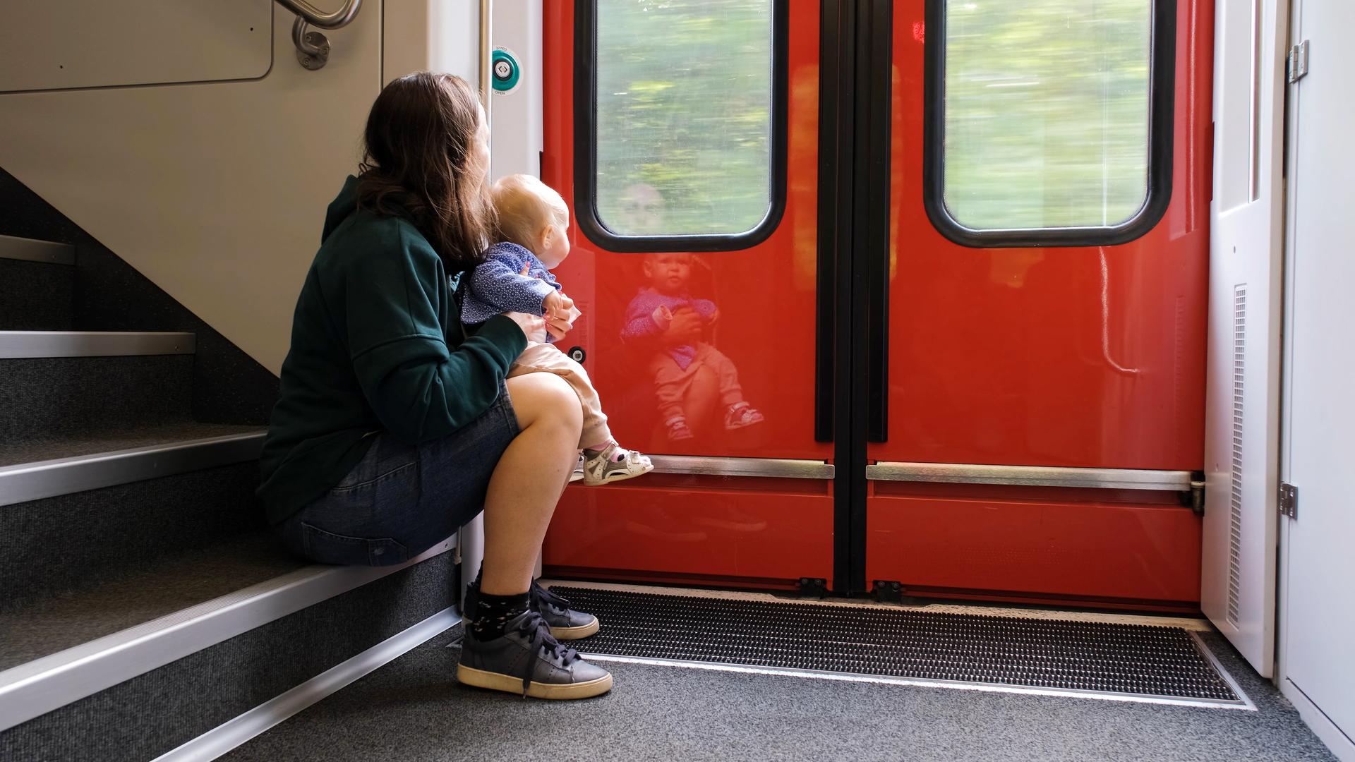 Eine Frau sitzt mit ihrem Kleinkind auf dem Schoß in einem Regionalzug auf der Treppe und schaut aus dem Fenster.