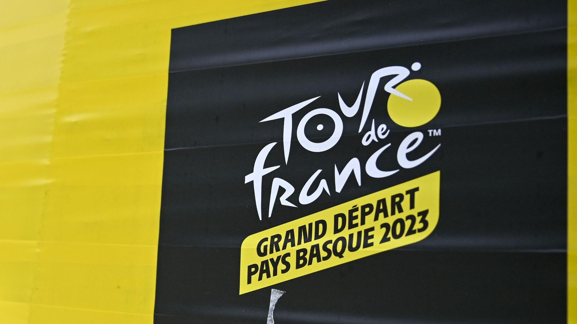 Auf dem Bild sieht man das Logo von der Tour de France auf einem gelben Unter-Grund.  Links davon stehen Bäume. 