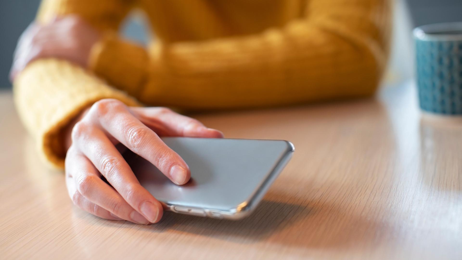 Eine Frau hält ein Smartphone in der Hand, das vor ihr auf dem Tisch liegt.