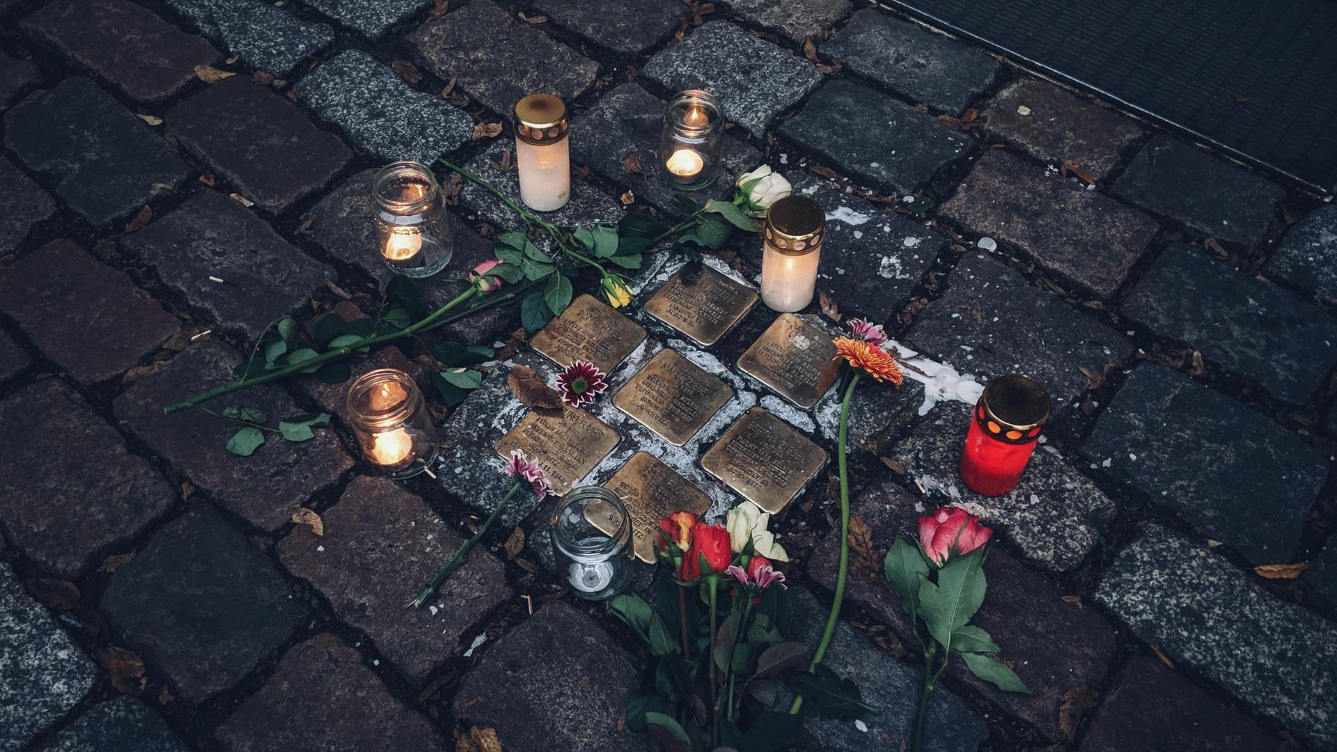 Kerzen und Blumen liegen um eine Ansammlung von sieben Stolpersteinen herum.