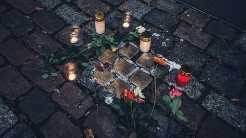 Kerzen und Blumen liegen um eine Ansammlung von sieben Stolpersteinen herum.