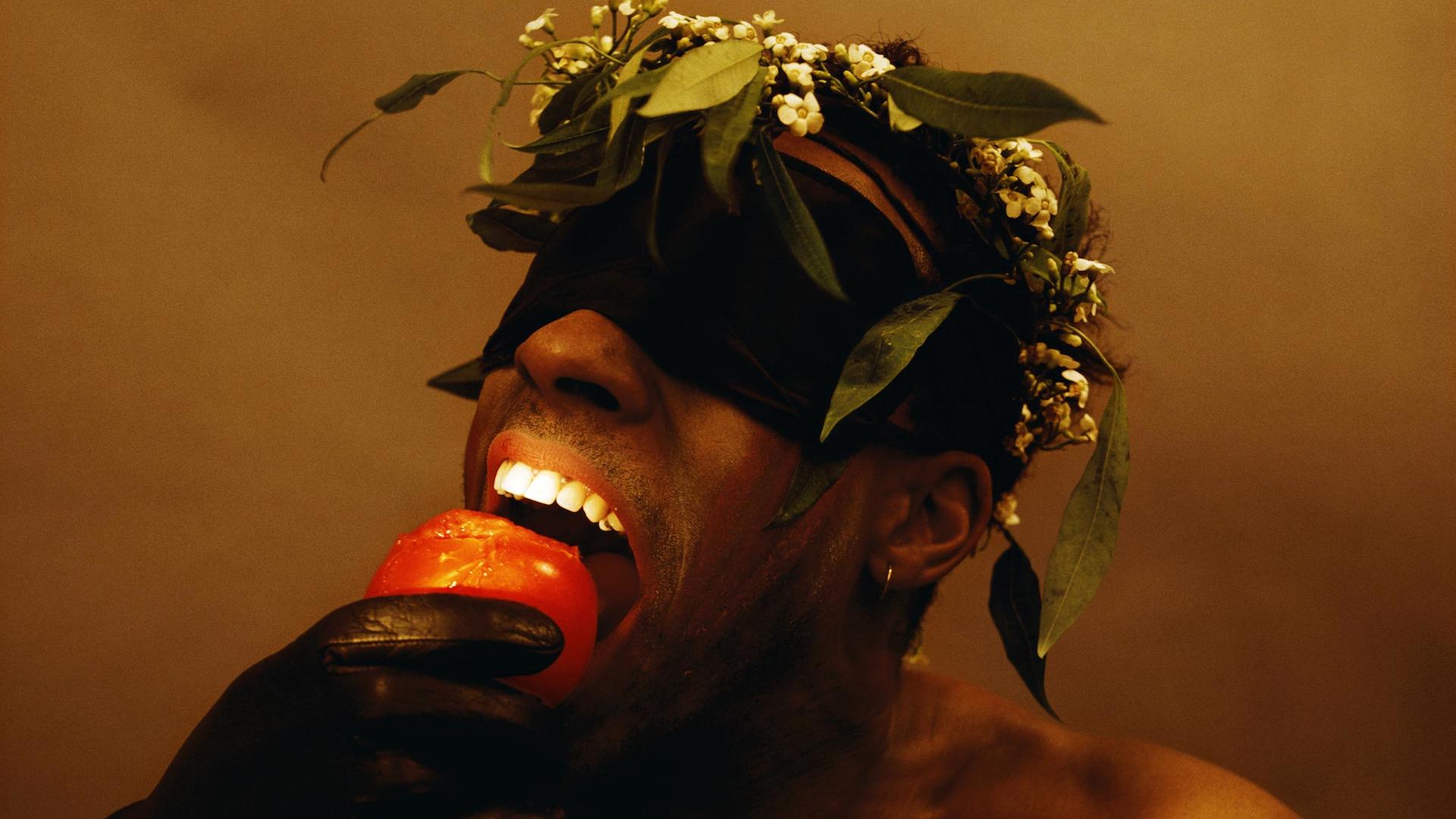 Ein Schwarzer mit Augenbinde und Lederhandschuhen beißt in eine Tomate. Er hat einen freien Oberkörper und trägt einen Blumenkranz auf dem Kopf.