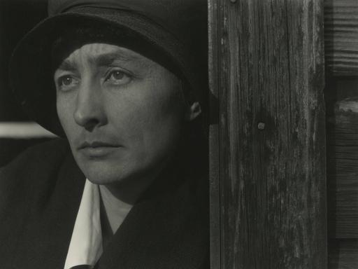 Schwarz-Weiß-Porträt von Georgia O’Keeffe mit Hut neben einer Holzwand, Blick seitlich aus dem Bild.