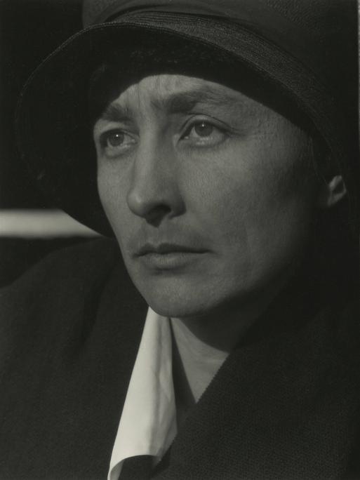 Schwarz-Weiß-Porträt von Georgia O’Keeffe mit Hut neben einer Holzwand, Blick seitlich aus dem Bild.