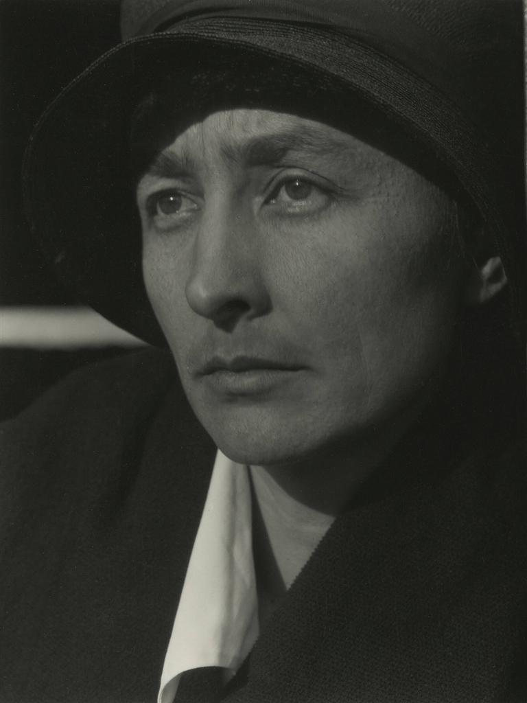 Schwarz-Weiß-Porträt von Georgia O’Keeffe mit Hut neben einer Holzwand, Blick seitlich aus dem Bild.