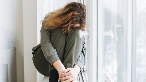 Die Schülerin berichtet von Depressionen und nächtlichen Panikgefühlen. Zu sehen: Eine junge Frau sitzt auf einer Fensterbank, hat den Kopf nach vorne gebeugt. Das Gesicht wird durch ihre langen Haare verdeckt.
