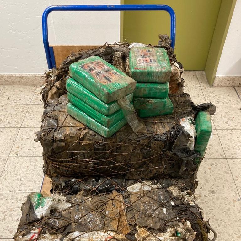 Der Transportwagen steht auf einem Fliesenboden. Auf zwei mit Netzen umwickelten Paketen stehen mehrere grün umwickelte kleinere Kokainpäckhen. Der Transportwagen steht auf einem Fliesenboden. Auf zwei mit Netzen umwickelten Paketen stehen mehrere grün umwickelte kleinere Kokainpäckhen.