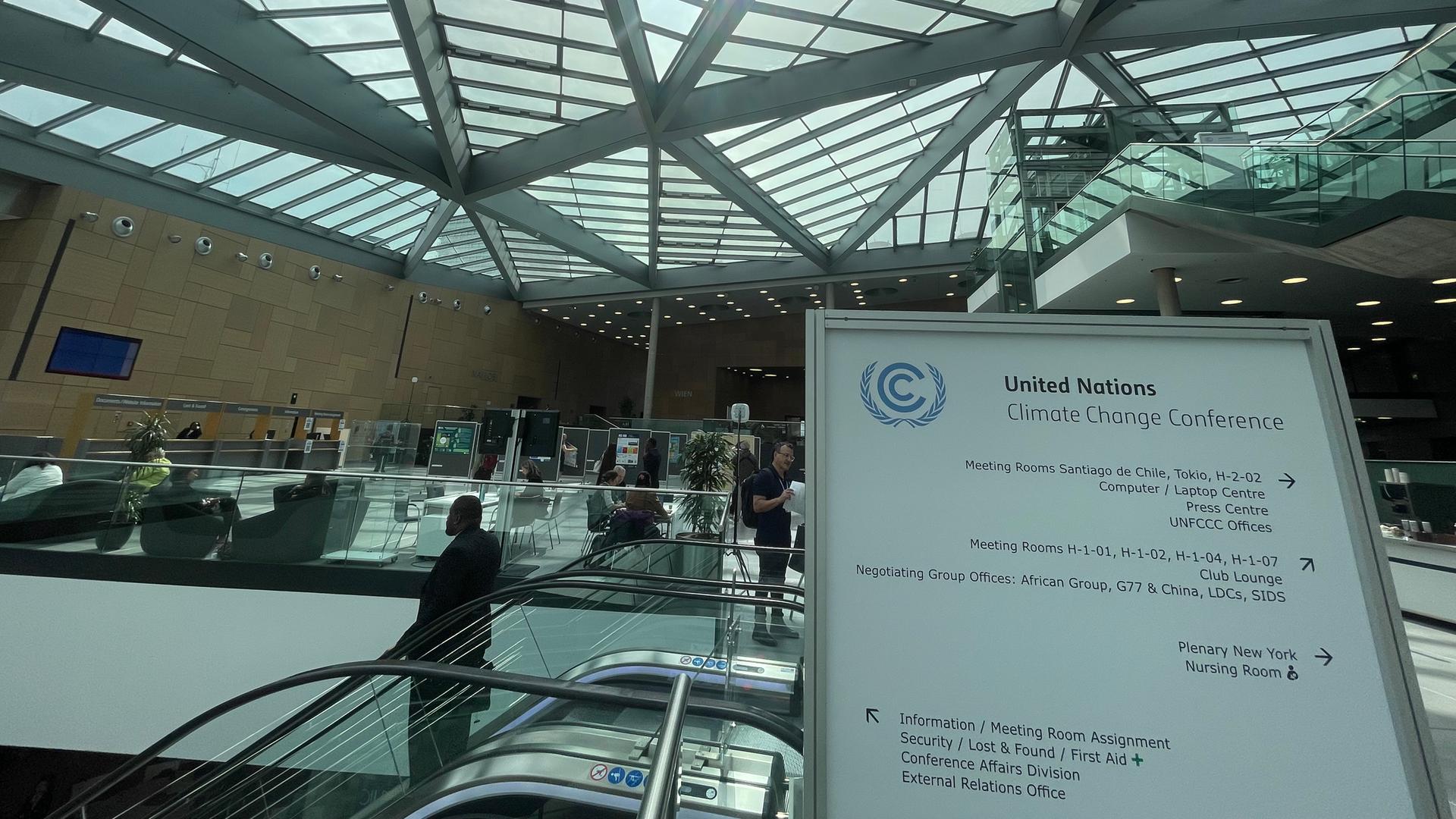 Ein Hinweisschild der UNO-Klimakonferenz steht im Foyer des World Conference Center in Bonn. Danben ist eine Rolltreppe auf der ein Mann nach unten fährt. Ein Hinweisschild der UNO-Klimakonferenz steht im Foyer des World Conference Center in Bonn. Danben ist eine Rolltreppe auf der ein Mann nach unten fährt.
