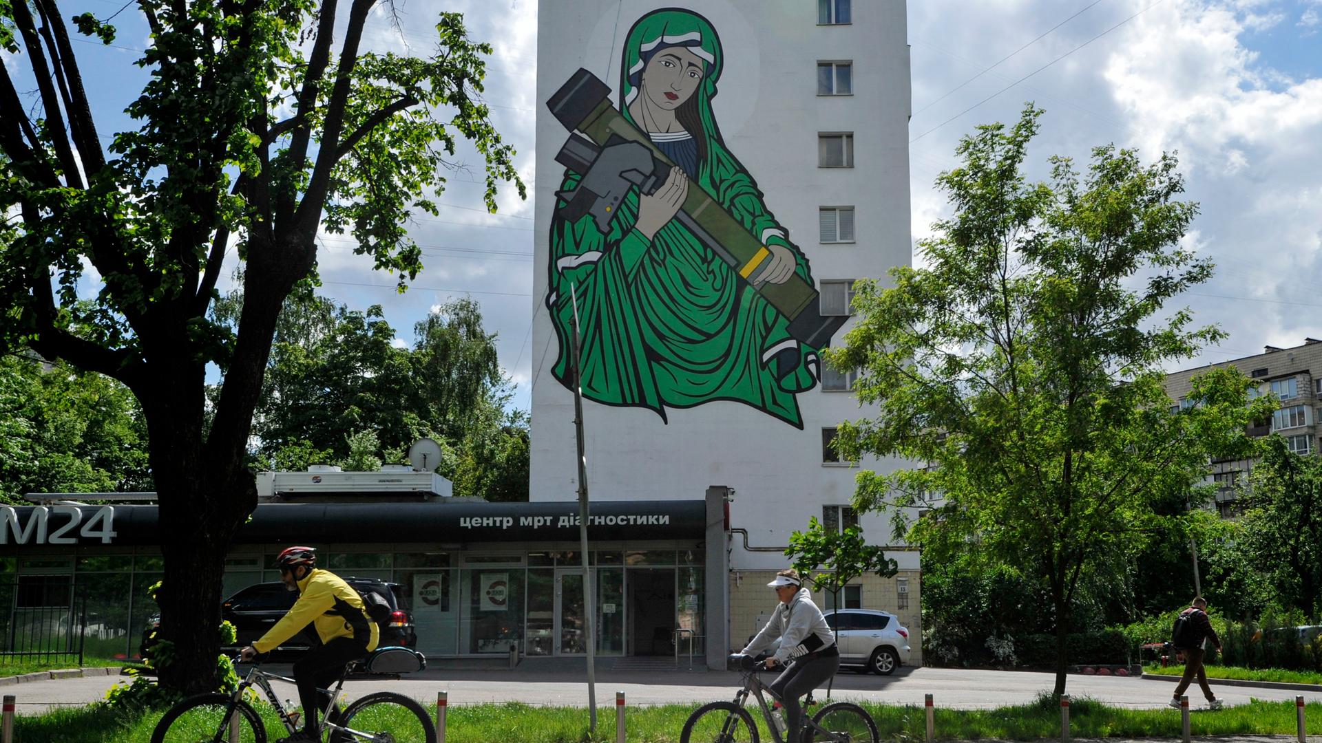 Radfahrer fahren an einem Wandgemälde vorbei, das die heilige Maria mit einer Panzerabwehrlenkwaffe in den Händen zeigt.