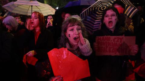 Demonstrantinnen in Warschau rufen und halten Schilder. Es regnet und sie tragen Regenschirme.