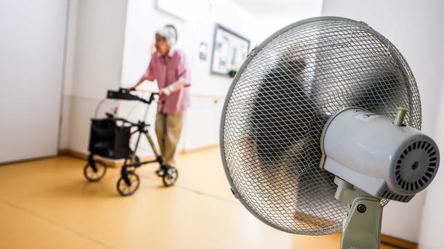 Eine Frau geht mit einem Rollator in einem Senioren-Heim über einen Flur, der aufgrund hoher Temperaturen mit einem Ventilator belüftet wird.