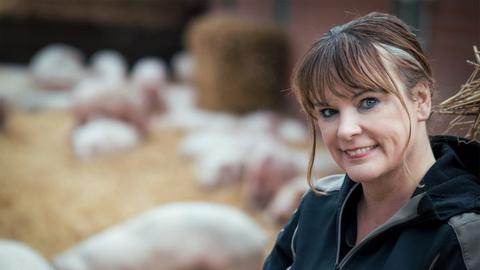 Gabriele Mörixmann trägt eine Wetterjacke und blickt freundlich in die Kamera. Im Hintergrund: Ferkel im Auslauf.