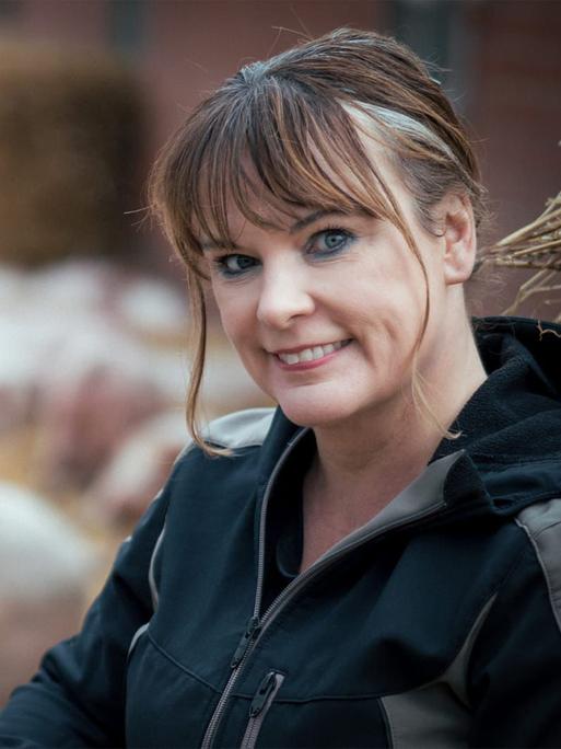 Gabriele Mörixmann trägt eine Wetterjacke und blickt freundlich in die Kamera. Im Hintergrund: Ferkel im Auslauf.