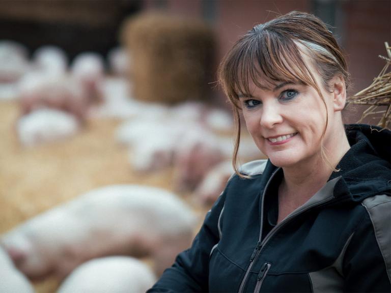 Gabriele Mörixmann trägt eine Wetterjacke und blickt freundlich in die Kamera. Im Hintergrund: Ferkel im Auslauf.