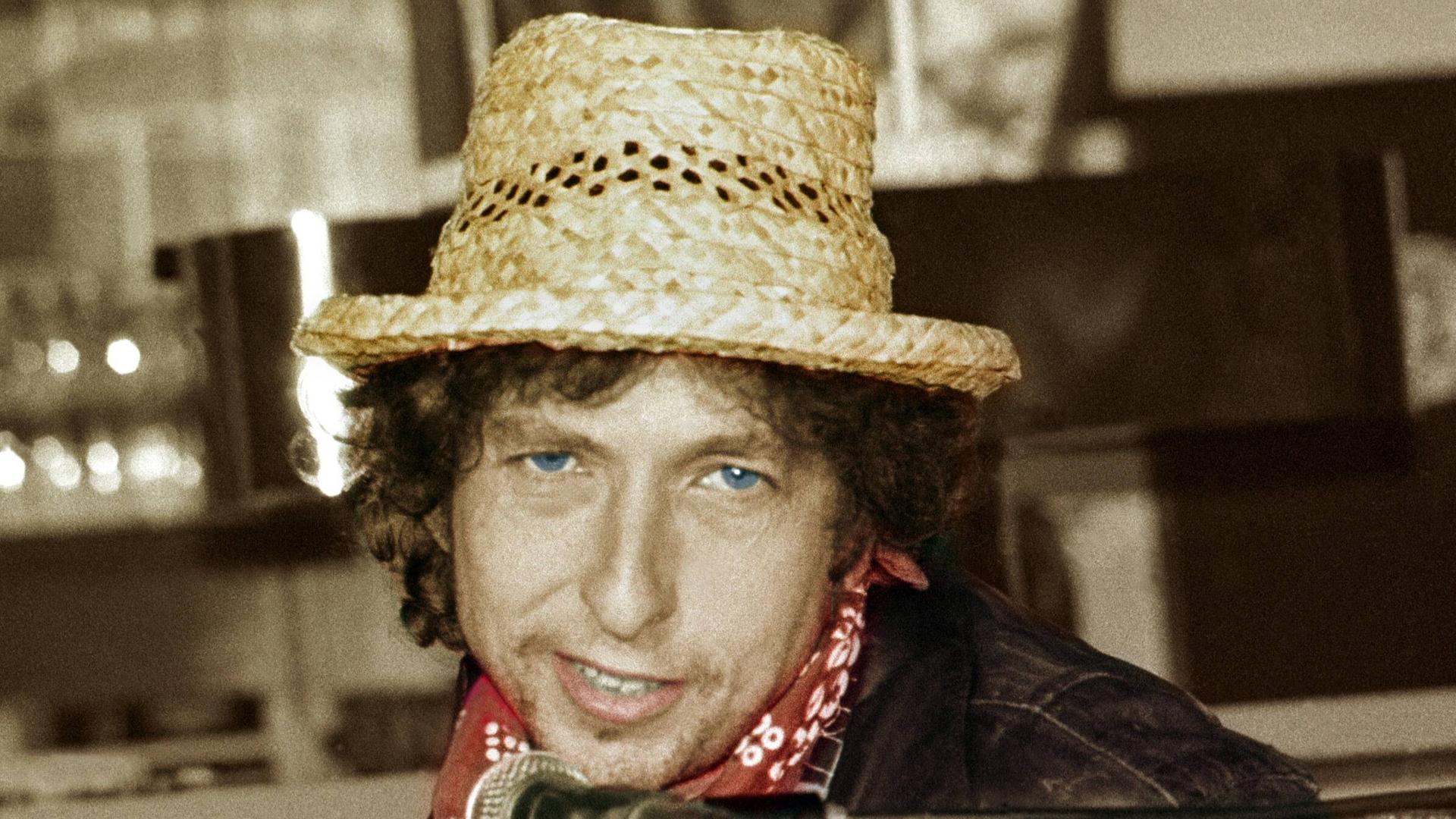 Ein weißer Mann in Lederjacke und mit blauen Augen, rotem Halstuch und Strohhut blickt in die Kamera. Unter seinem Kinn hängt ein Mikrofon. Es ist Bob Dylan auf einem Foto von 1987.