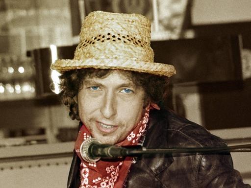 Ein weißer Mann in Lederjacke und mit blauen Augen, rotem Halstuch und Strohhut blickt in die Kamera. Unter seinem Kinn hängt ein Mikrofon. Es ist Bob Dylan auf einem Foto von 1987.