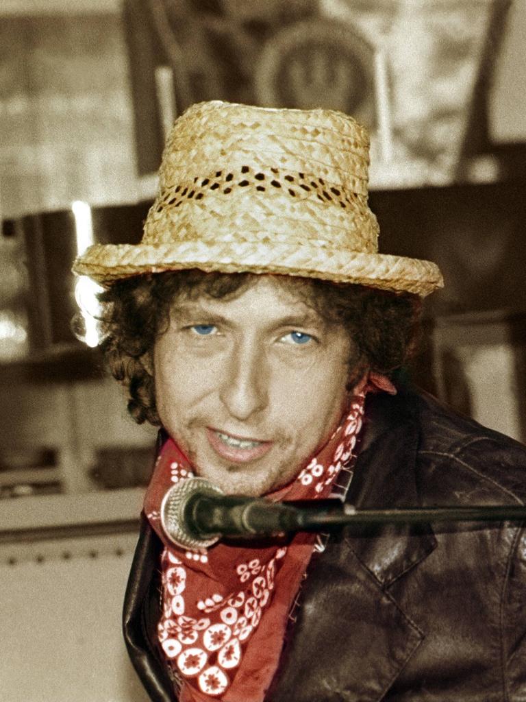 Ein weißer Mann in Lederjacke und mit blauen Augen, rotem Halstuch und Strohhut blickt in die Kamera. Unter seinem Kinn hängt ein Mikrofon. Es ist Bob Dylan auf einem Foto von 1987.