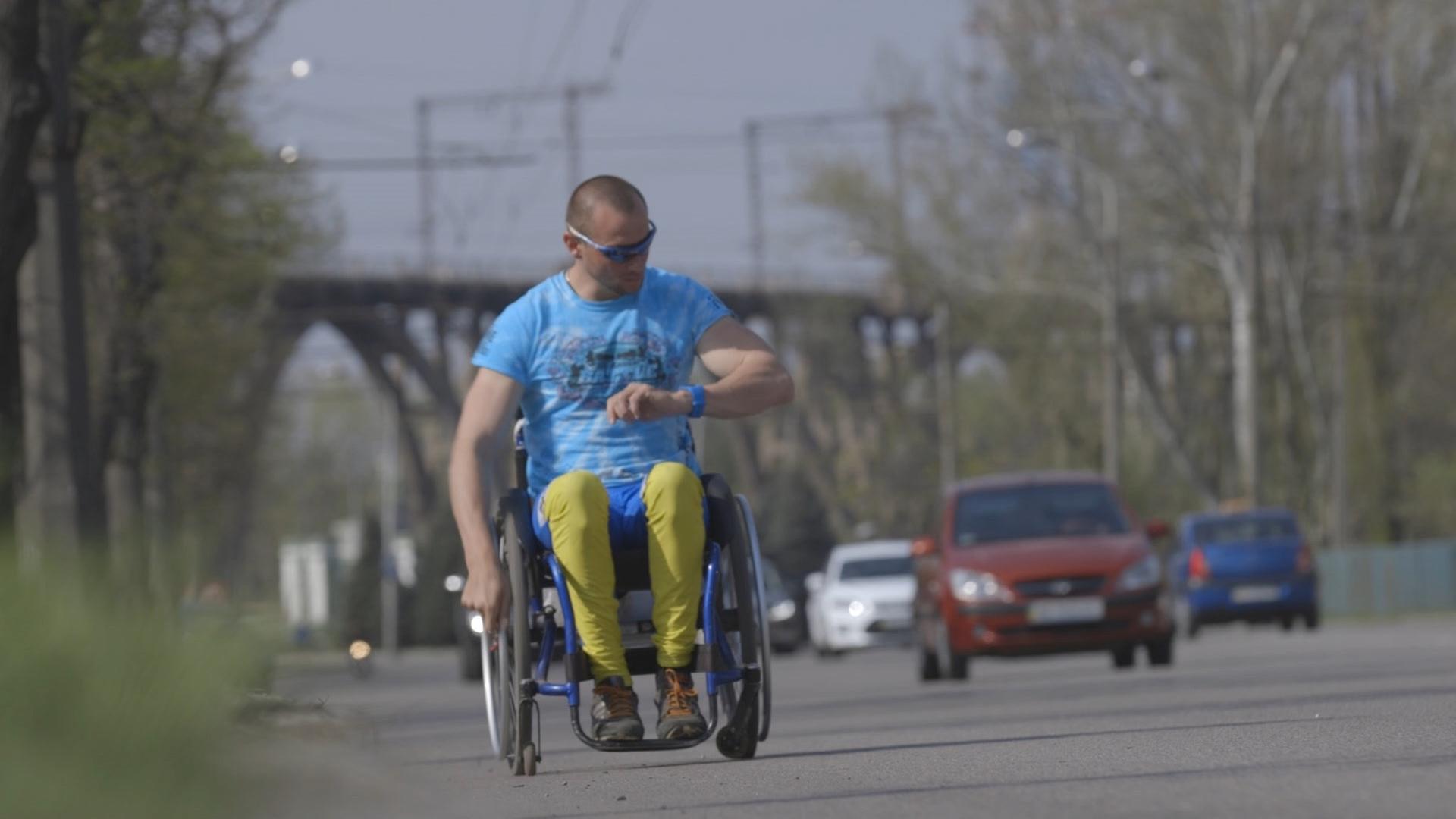 Ein Rollstuhlfahrer fährt am Rand einer befahrenen Straße und blickt auf seine Uhr: Szene aus dem Film "Pushing Boundaries" von Lesia Cordonets.