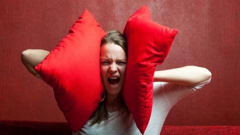 Eine Frau hält sich zwei rote Samtkissen auf die Ohren und hat den Mund weit geöffnet. Sie scheint gegen den Lärm anzuschreien. 