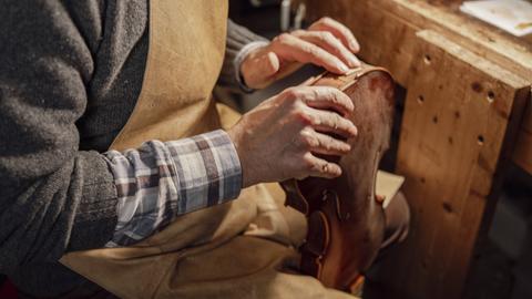 Geigenbauer in einer Werkstatt mit Schürze und Karohemd untersucht mit seinen Händen den Körper einer Geige