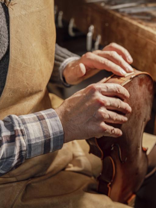Geigenbauer in einer Werkstatt mit Schürze und Karohemd untersucht mit seinen Händen den Körper einer Geige