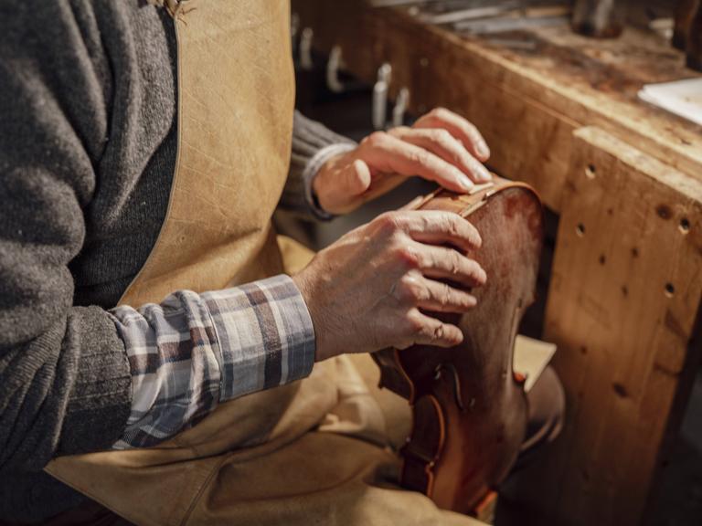 Geigenbauer in einer Werkstatt mit Schürze und Karohemd untersucht mit seinen Händen den Körper einer Geige