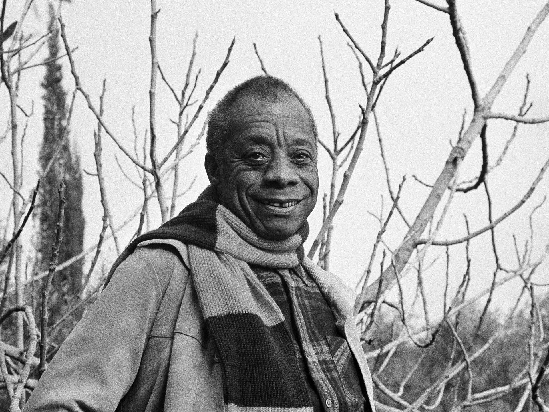 Der US-amerikanische Autor James Baldwin auf einer alten Schwarz-weiß-Aufnahme. Er trägt einen Schal und lacht vergnügt. 