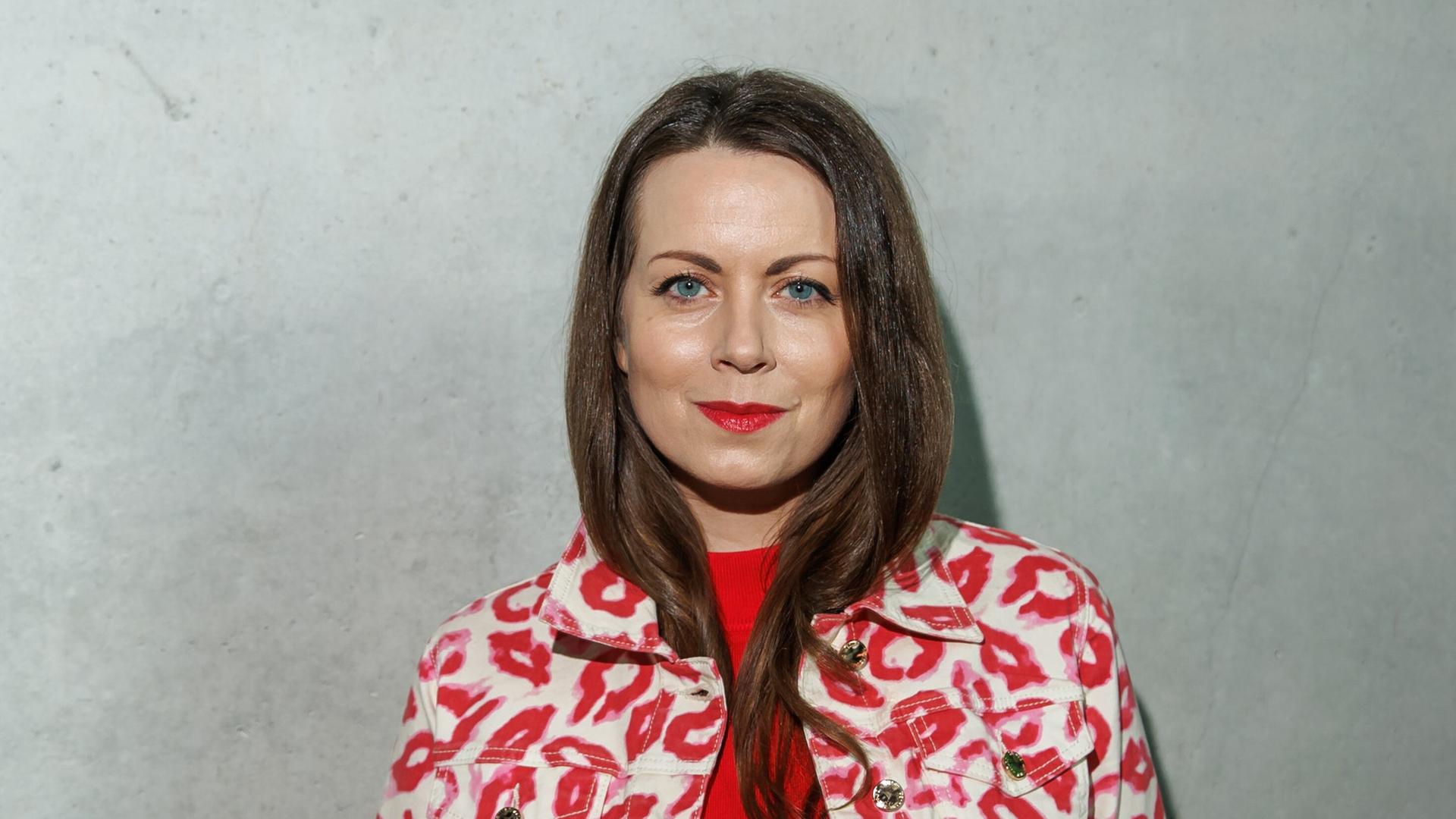 Alice Dwyer trägt eine rote Hose, ein rotes Oberteil und eine weißrot gemusterte Jacke. Sie steht vor einer grauen Wand und lächelt freundlich in die Kamera. Alice Dwyer trägt eine rote Hose, ein rotes Oberteil und eine weißrot gemusterte Jacke. Sie steht vor einer grauen Wand und lächelt freundlich in die Kamera.