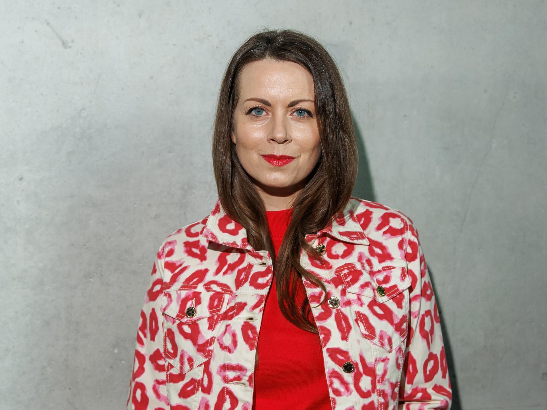 Alice Dwyer trägt eine rote Hose, ein rotes Oberteil und eine weißrot gemusterte Jacke. Sie steht vor einer grauen Wand und lächelt freundlich in die Kamera.