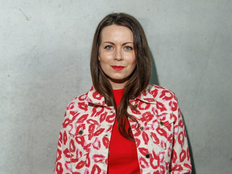 Alice Dwyer trägt eine rote Hose, ein rotes Oberteil und eine weißrot gemusterte Jacke. Sie steht vor einer grauen Wand und lächelt freundlich in die Kamera.