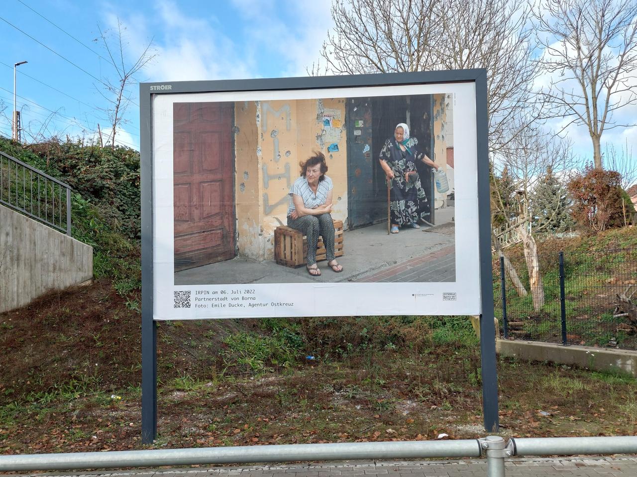 Das Foto auf einer Plakatwand zeigt zwei Frauen for ihrem zerstörten Haus.