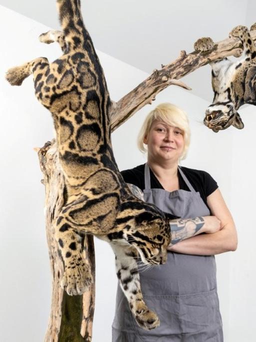 Die Tierpräparatorin Christin Scheinpflug, in blauer Arbeitsschürze und selbstbewusst verschränkten Armen, steht zwischen zwei ausgestopften Leoparden, die an einem Baumstamm hängen.