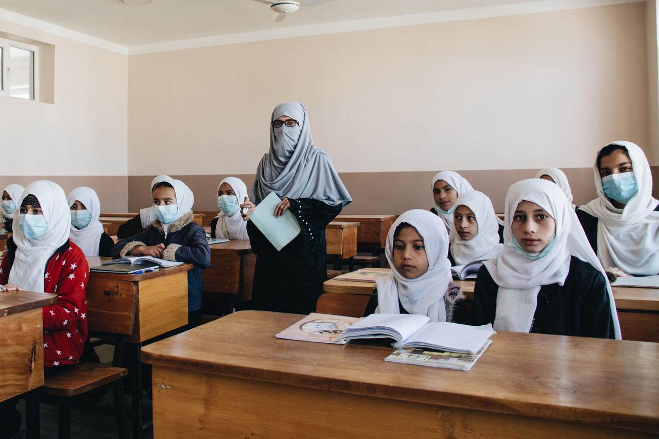 Schülerinnen mit Kopftuch sitzen an Pulten, eine Lehrerin mit Kopftuch steht im Klassenraum: die Safaa-Schule im Norden Kabuls, Afghanistan.