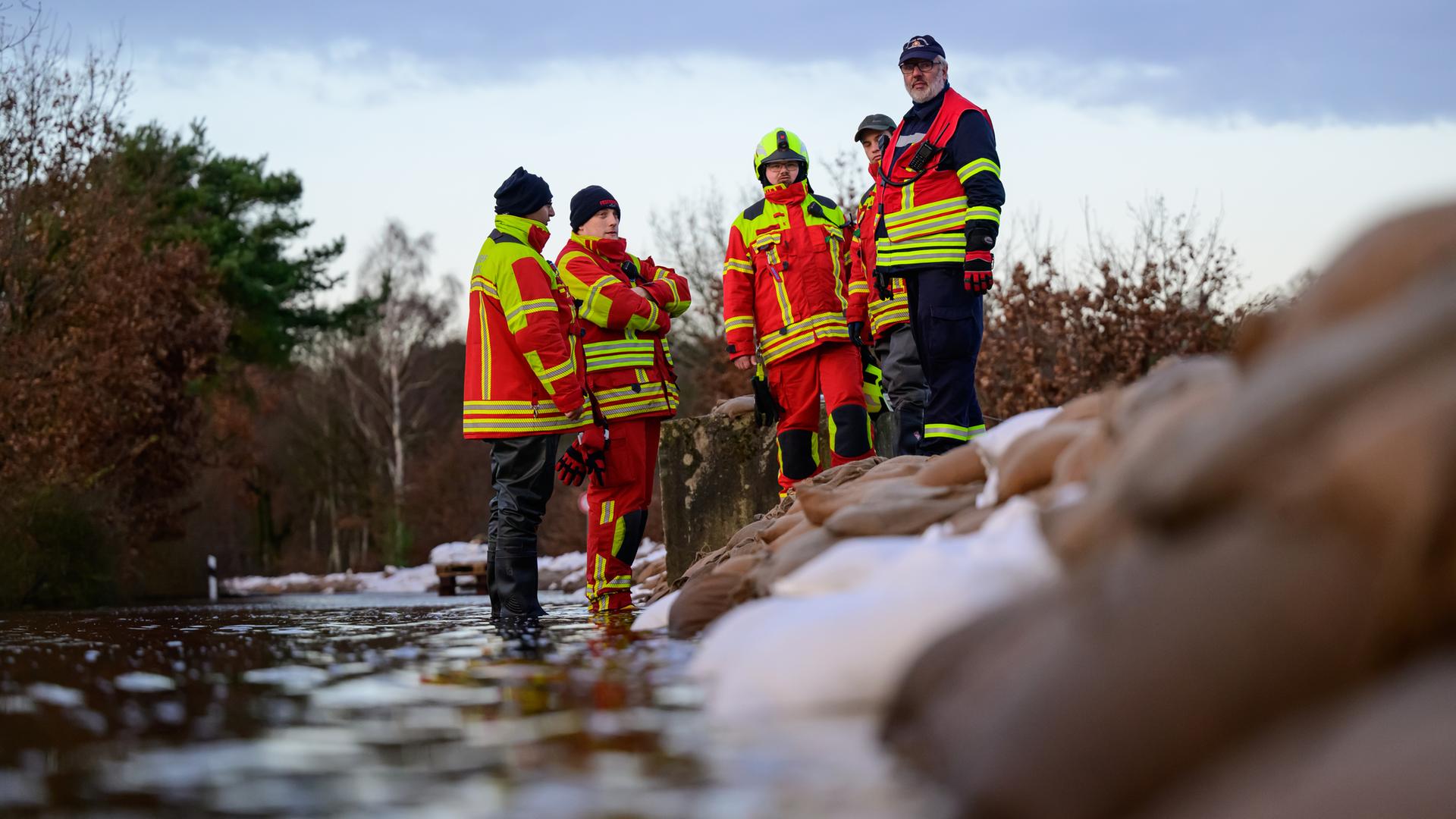 Mehrere ehrenamtliche Einsatzkräfte der Feuerwehr in Uniform stehen zur Besprechung in einer Gruppe zusammen neben aufeinandergeschichteten Sandsäcken.