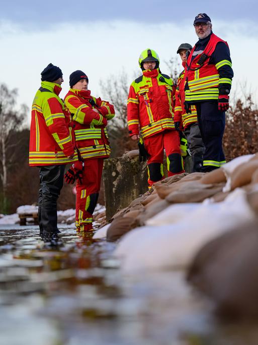 Mehrere ehrenamtliche Einsatzkräfte der Feuerwehr in Uniform stehen zur Besprechung in einer Gruppe zusammen neben aufeinandergeschichteten Sandsäcken.